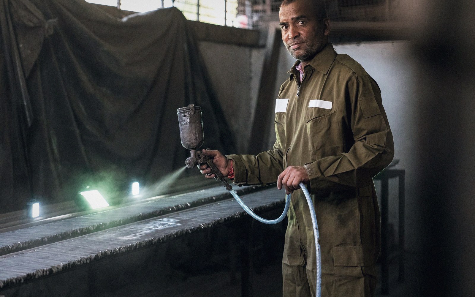 A worker in coveralls holds a spray gun beside a workshop conveyor bathed in green light