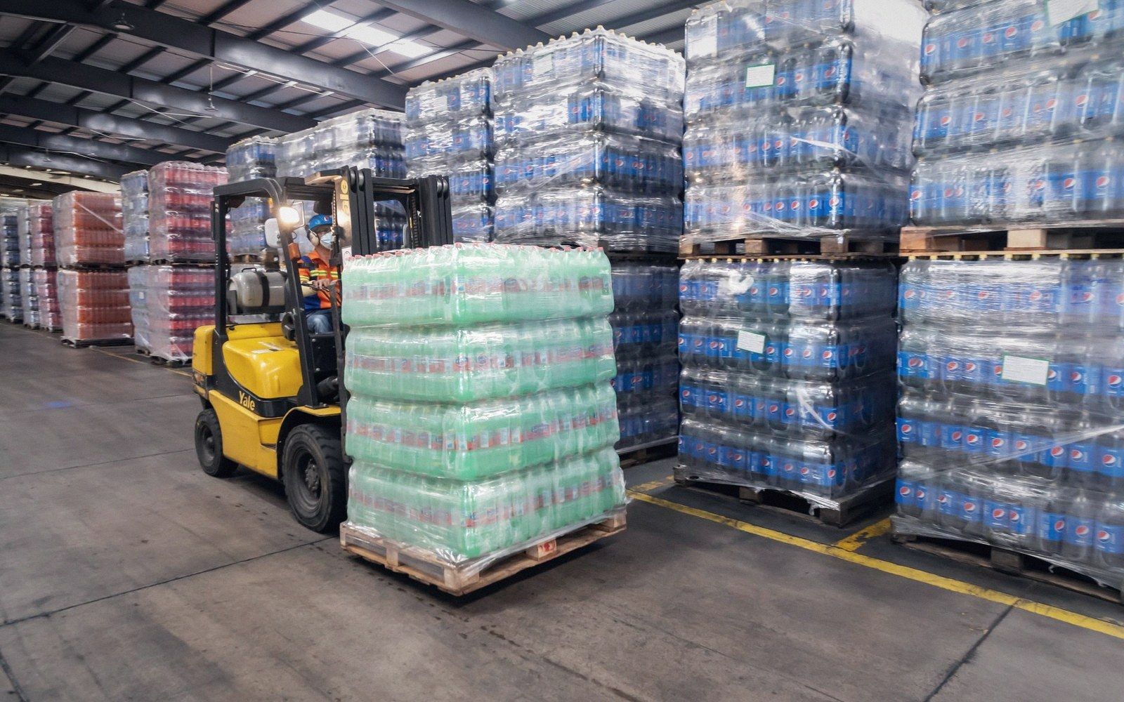 A forklift moves a shrink-wrapped pallet of bottled beverages through a warehouse aisle