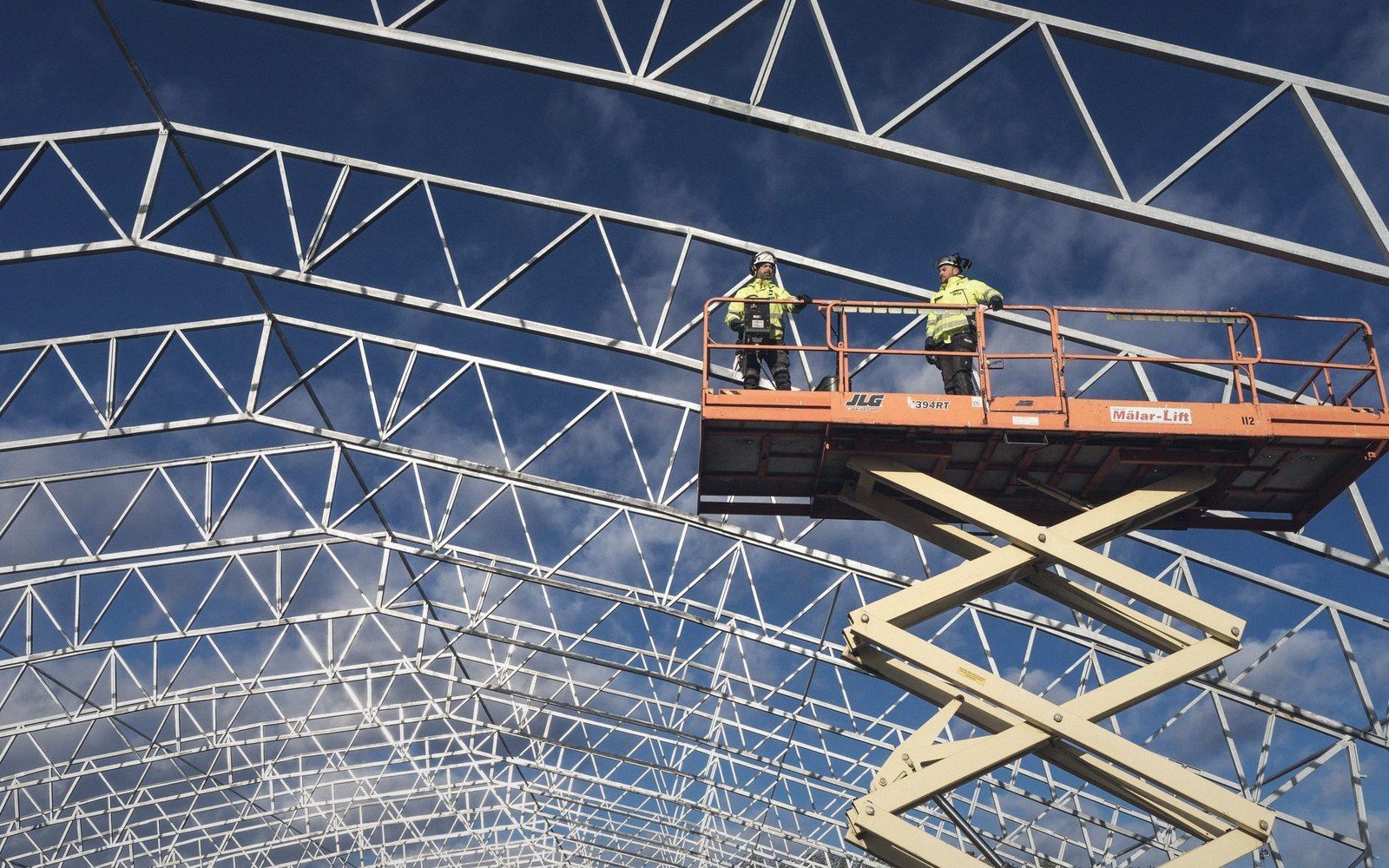 Two workers on a scissor lift inspect a triangulated steel roof truss structure