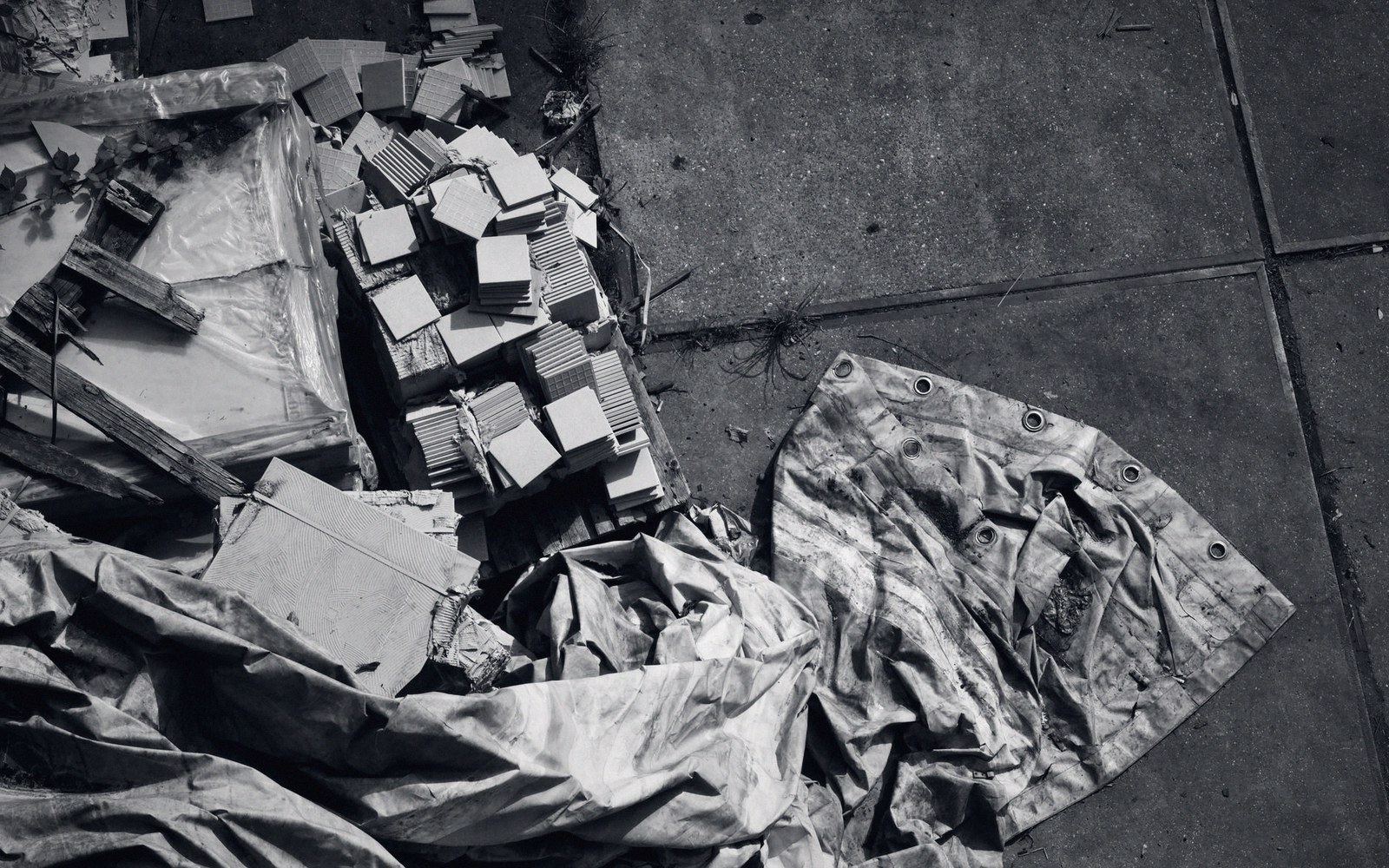 Stacked tiles and a crumpled tarp lie beside a torn canvas sheet on pavement